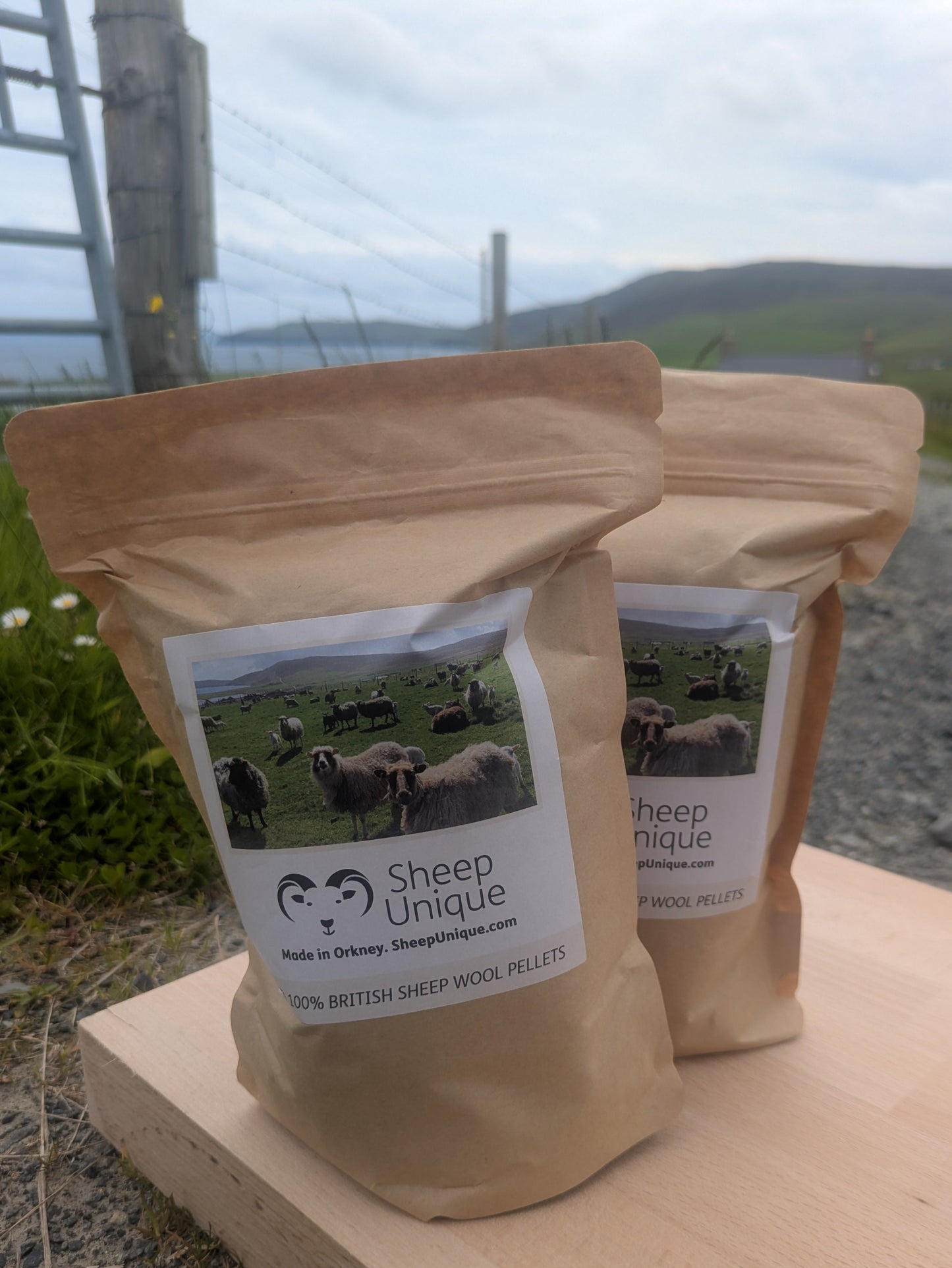 Two packages of Sheep Unique wool pellets on a wooden surface with a scenic background.