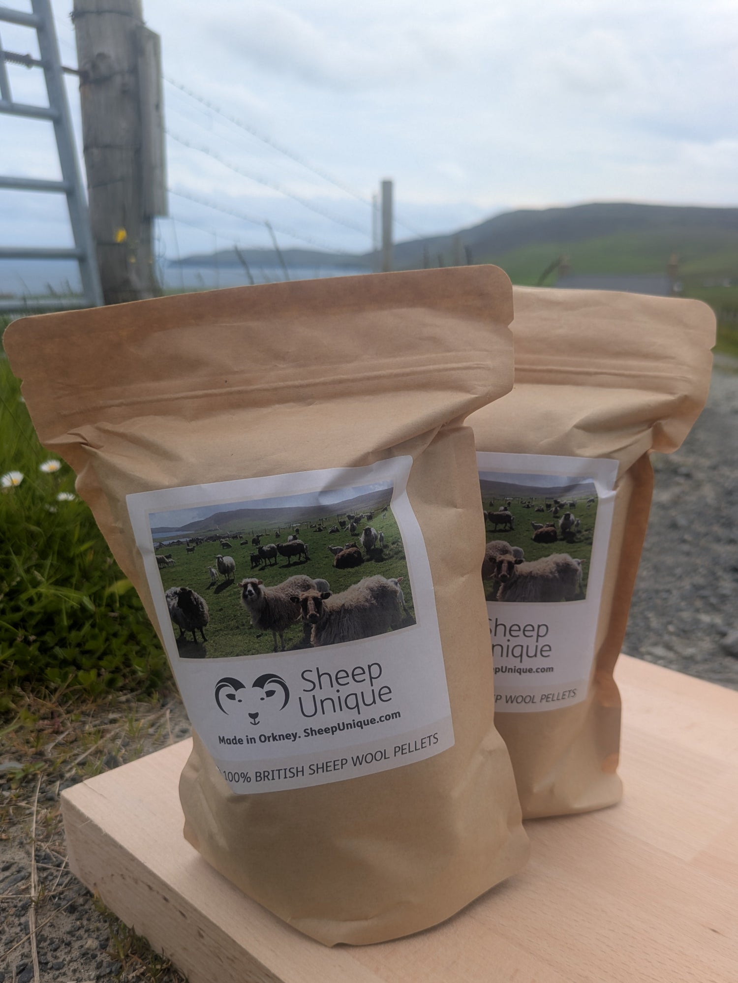 Two packages of Sheep Unique wool pellets on a wooden surface with a scenic background.